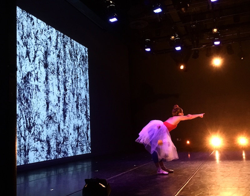 A woman in a tulle skirt bends at her waist and points her finger towards the audience